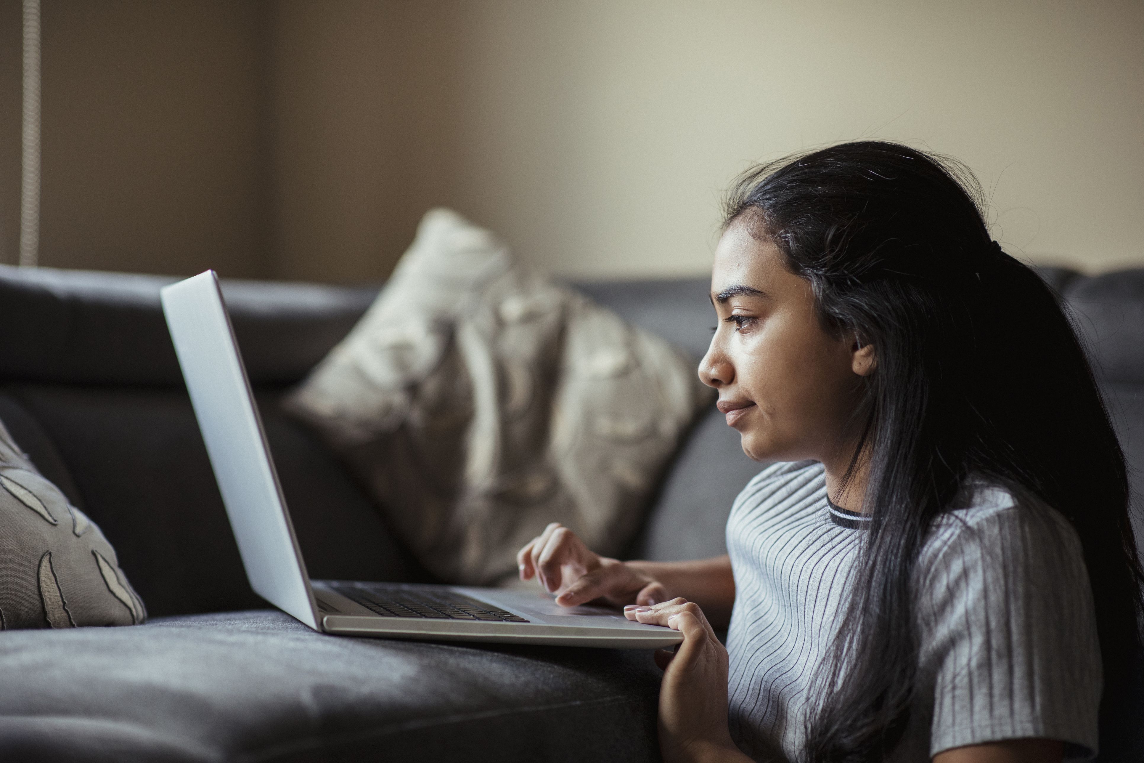 A young girl is kneeling on the floor in her living room, using her laptop that is sitting on the sofa.