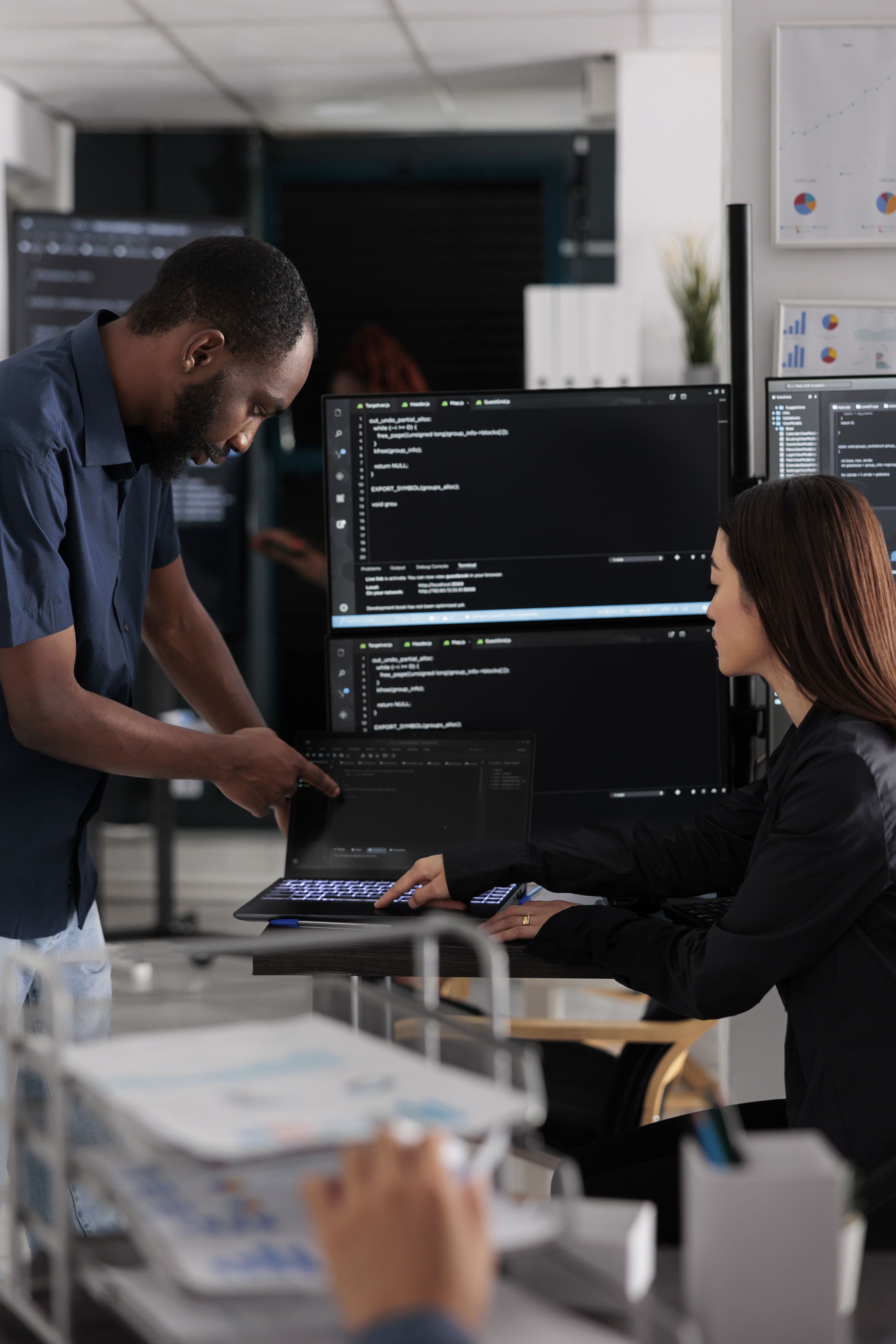 Image of a man and woman testing out a laptop. Behind them, a large monitor screen with code on it