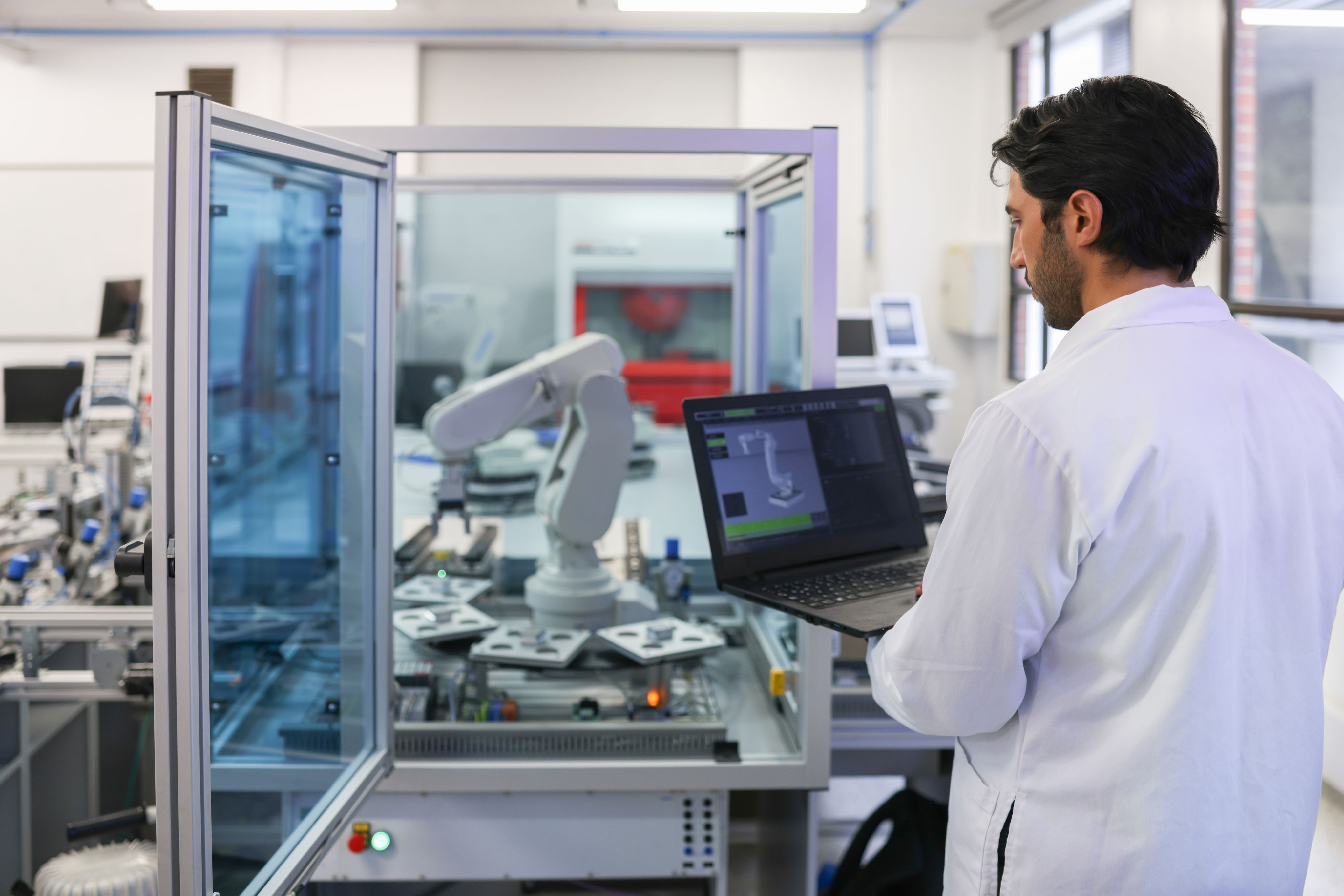 Man in a robotics lab holding a laptop with a model of the robotic arm on the table in front.