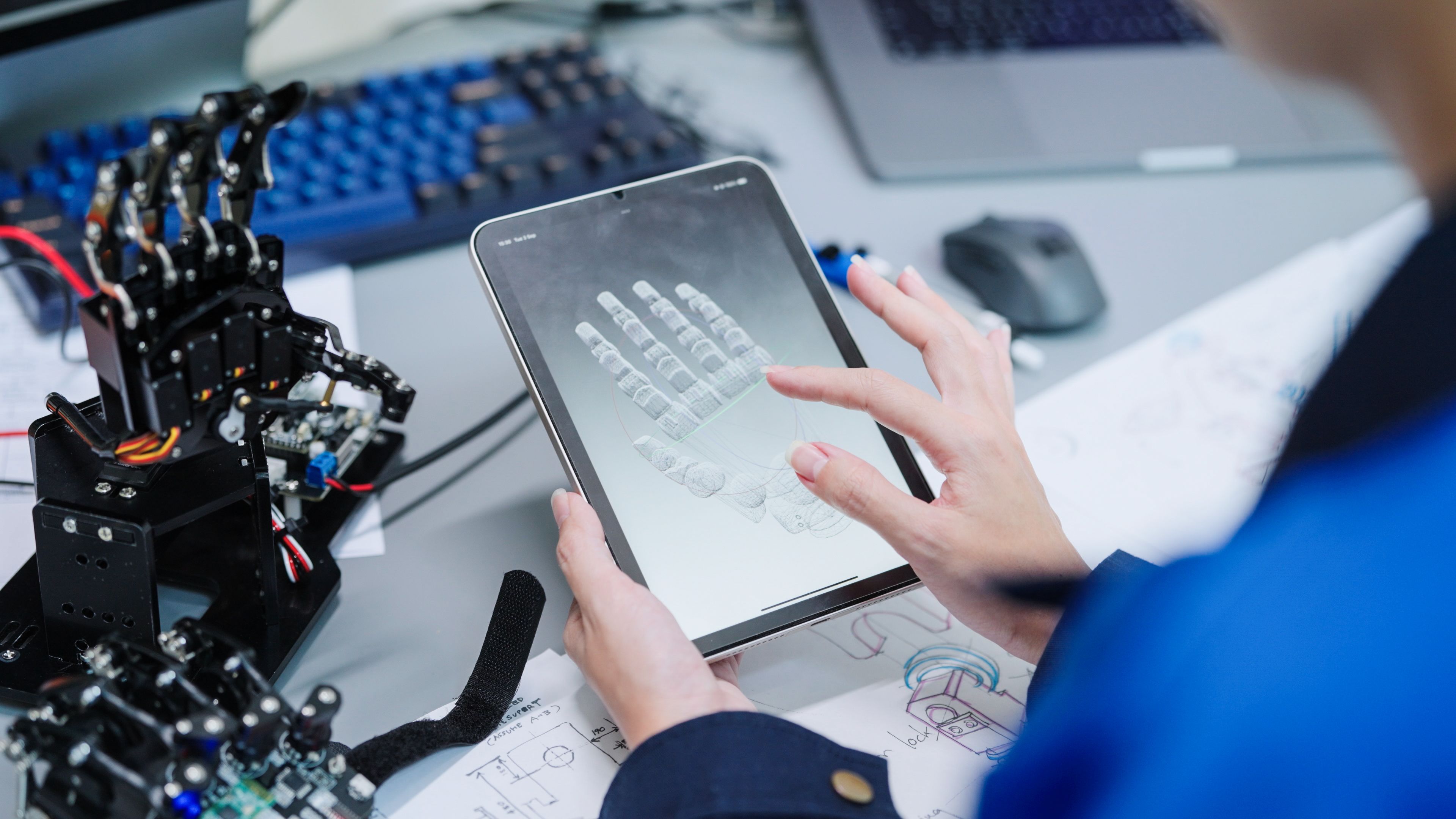 Person holding an IPad looking at a model of a robotic hand on their screen. Robotic hand sat on the desk next to them.