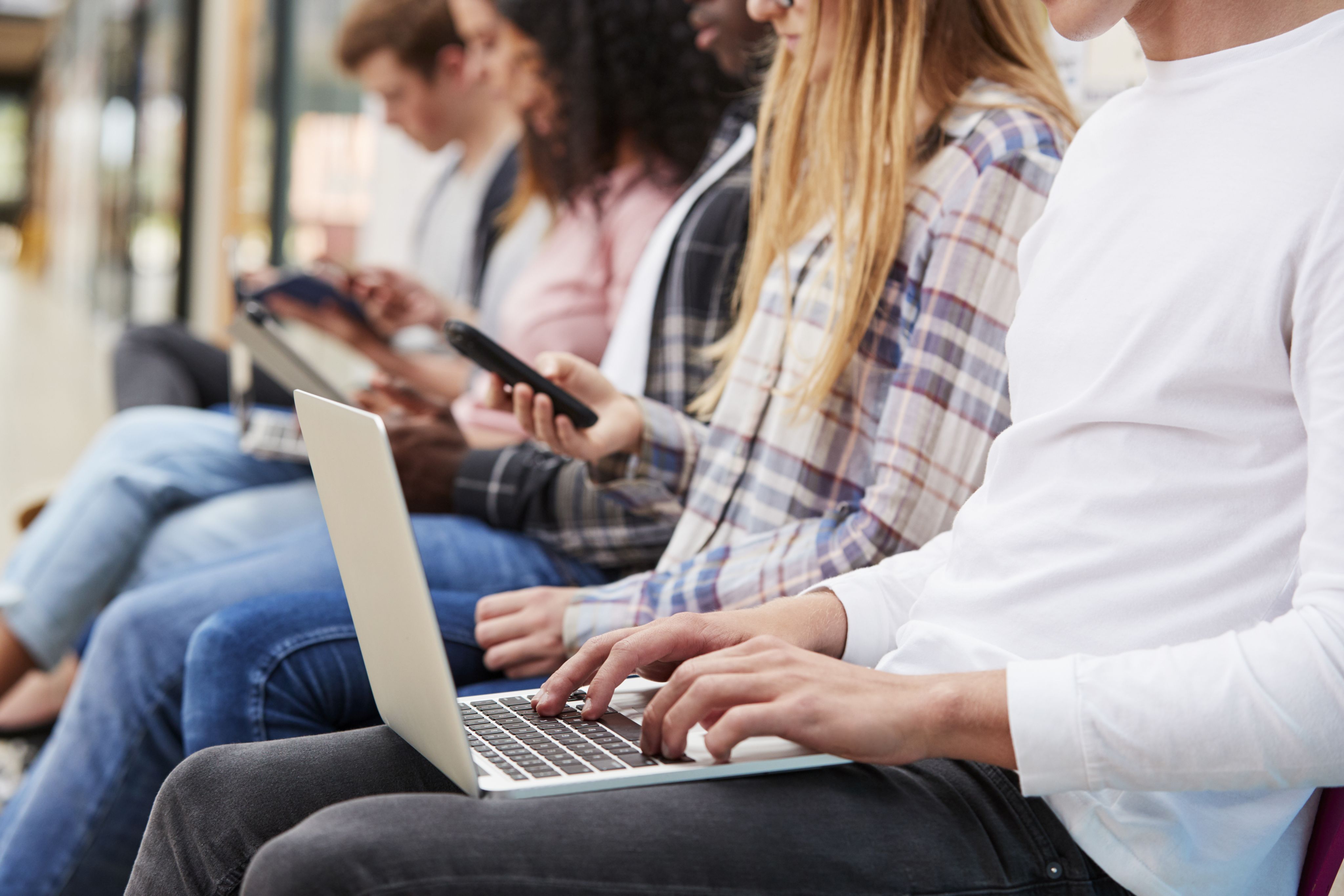 Young people sat in a row looking at their phones and working on laptops.