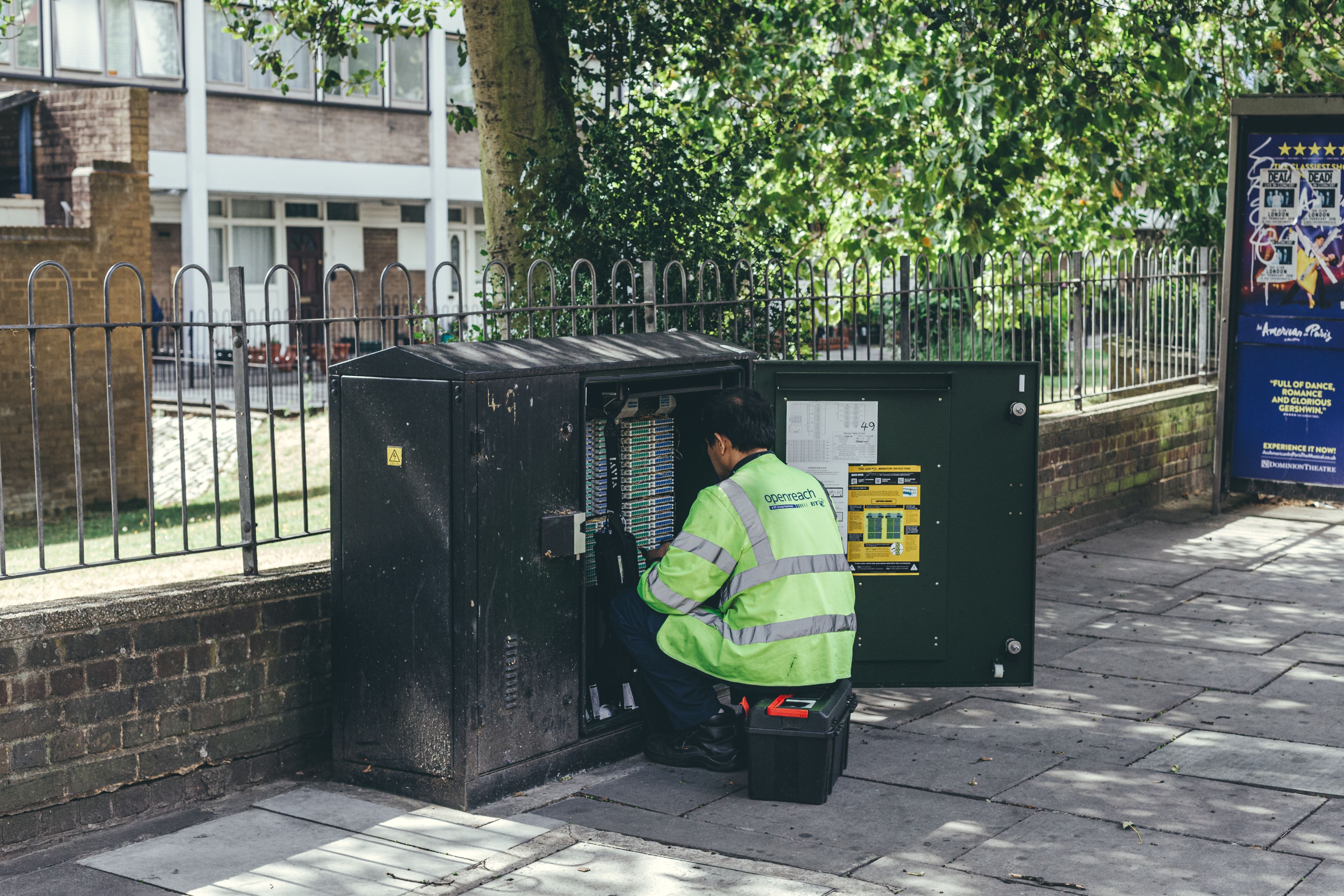 A technician in a yellow reflective jacket works on an open telecommunications cabinet on a pavement, with trees and residential buildings in the background.
