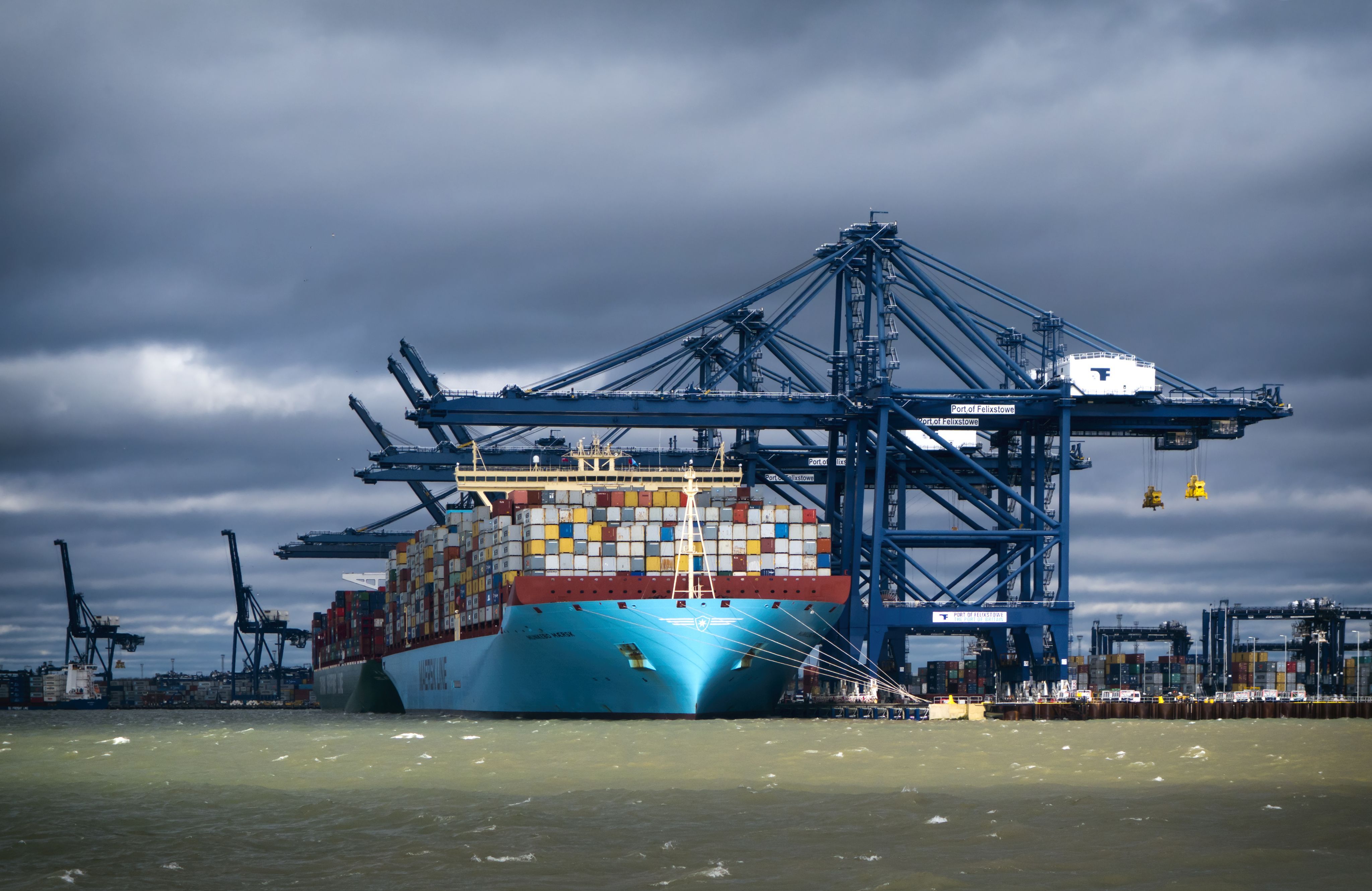 A container ship docked at a port with blue cranes and multicoloured containers under stormy skies.