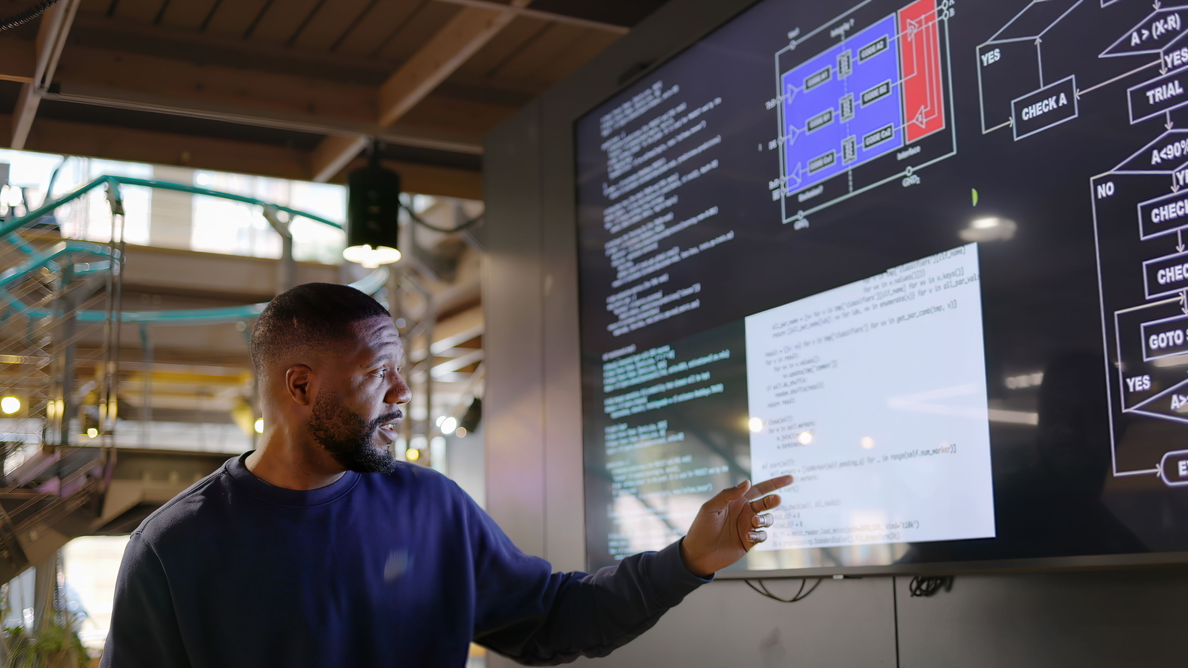 man is pointing at a large screen displaying technical diagrams and code, likely explaining or presenting the content in a modern office or tech environment.