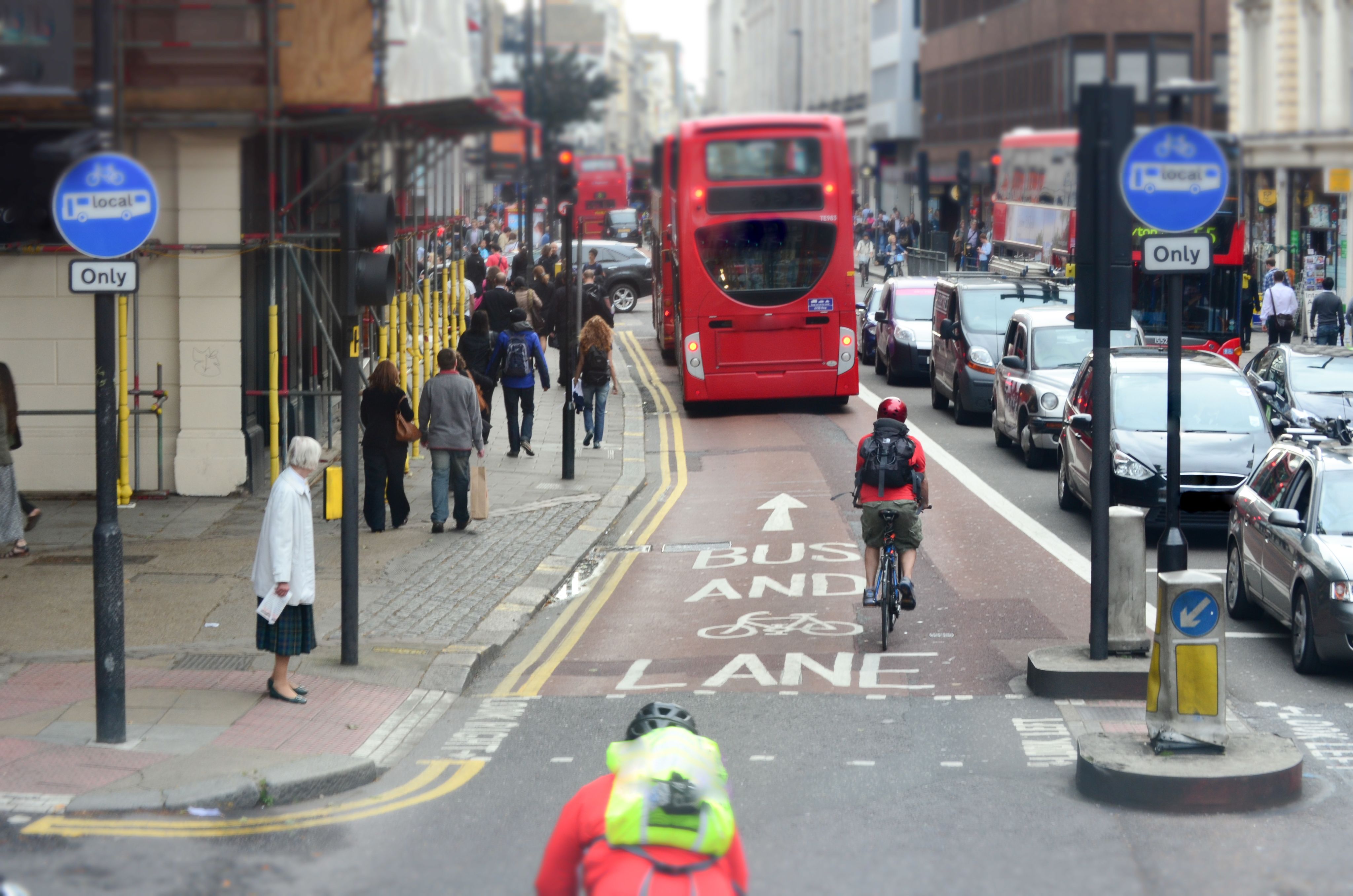 a busy urban street with a red double-decker bus in the bus lane, a cyclist riding behind it, pedestrians walking on the sidewalk, and cars lined up in traffic on the adjacent lane.