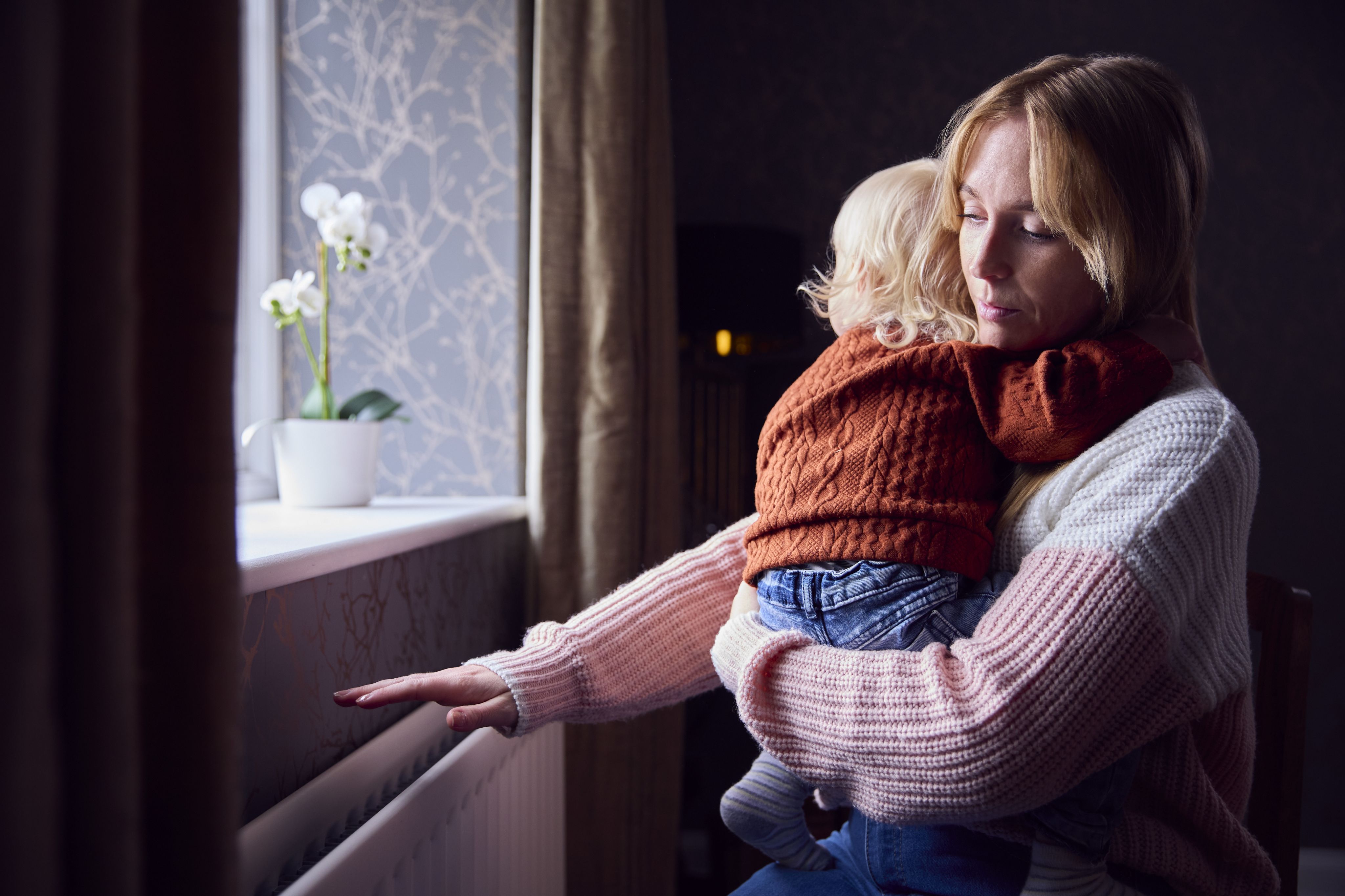 Mother With Son Trying To Keep Warm By Radiator At Home During Cost Of Living Energy Crisis.