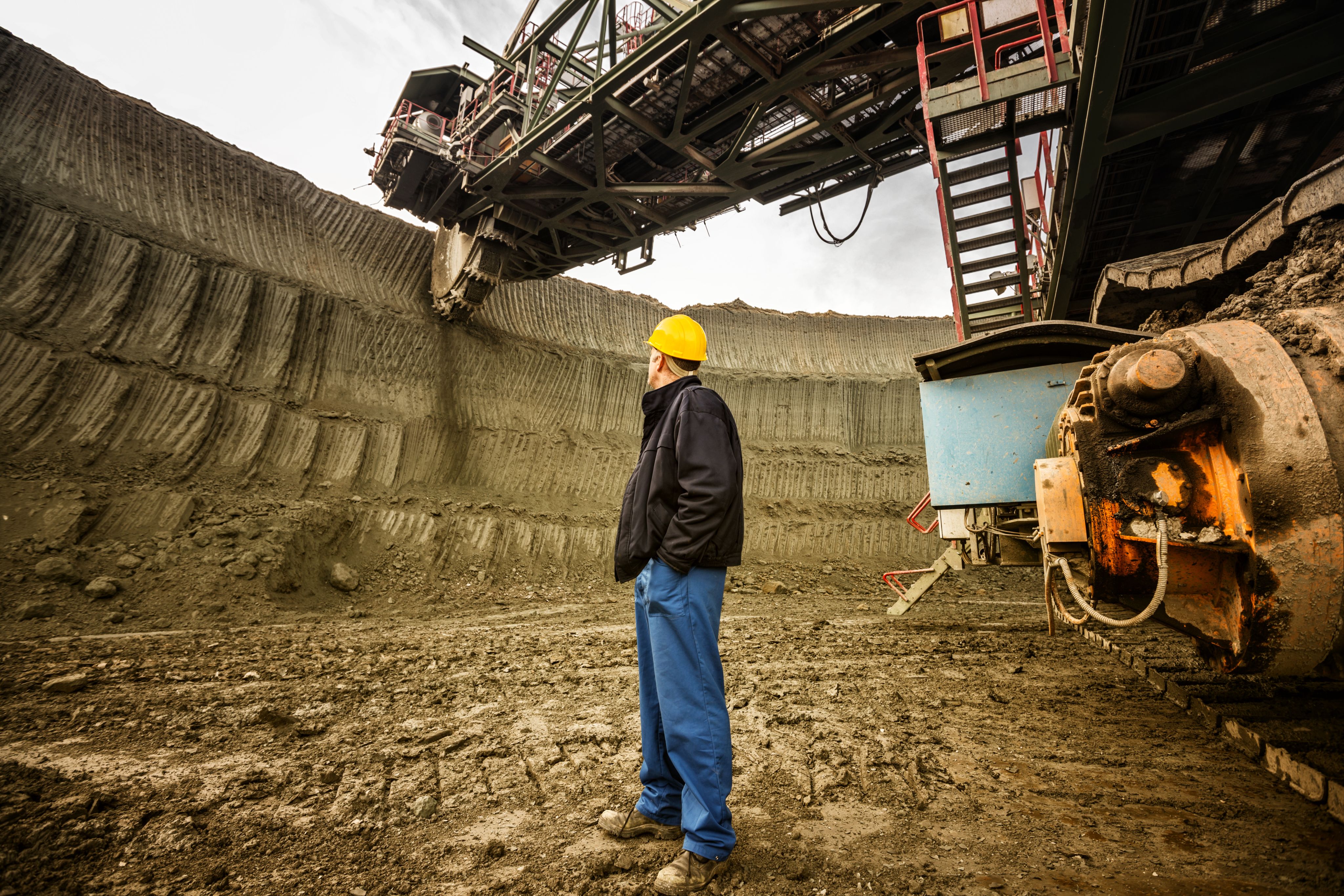 Coal mine worker with a helmet on his head standing in front of huge drill machine and looking at it. Rear view.