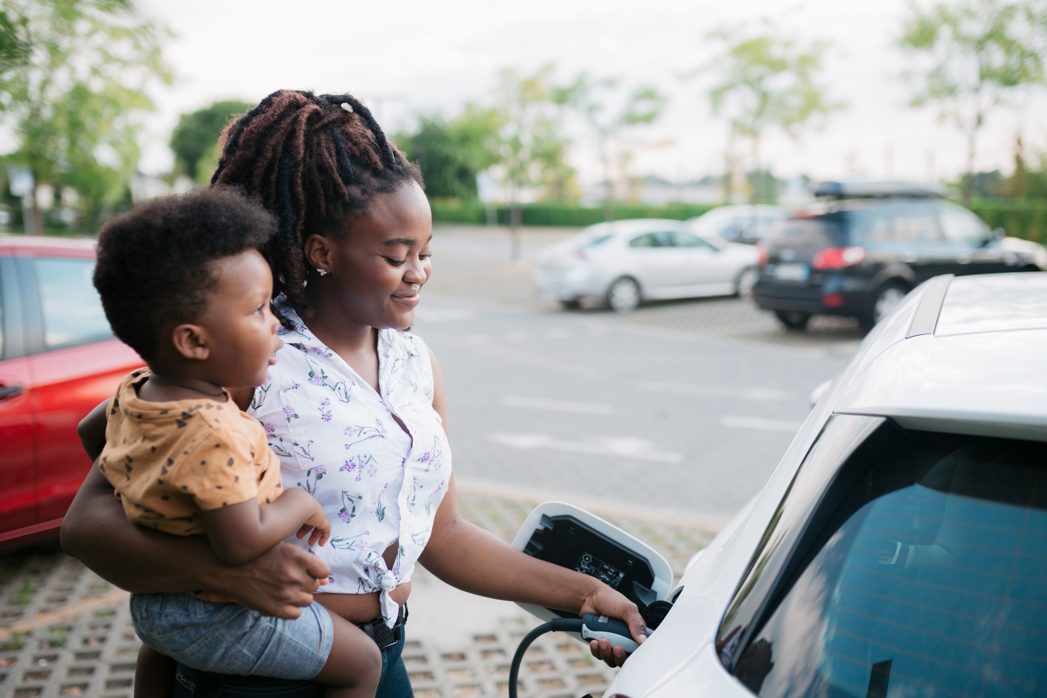 Young adult mother charging her electric car parked next to the charging station and holding her baby son in her other hand.