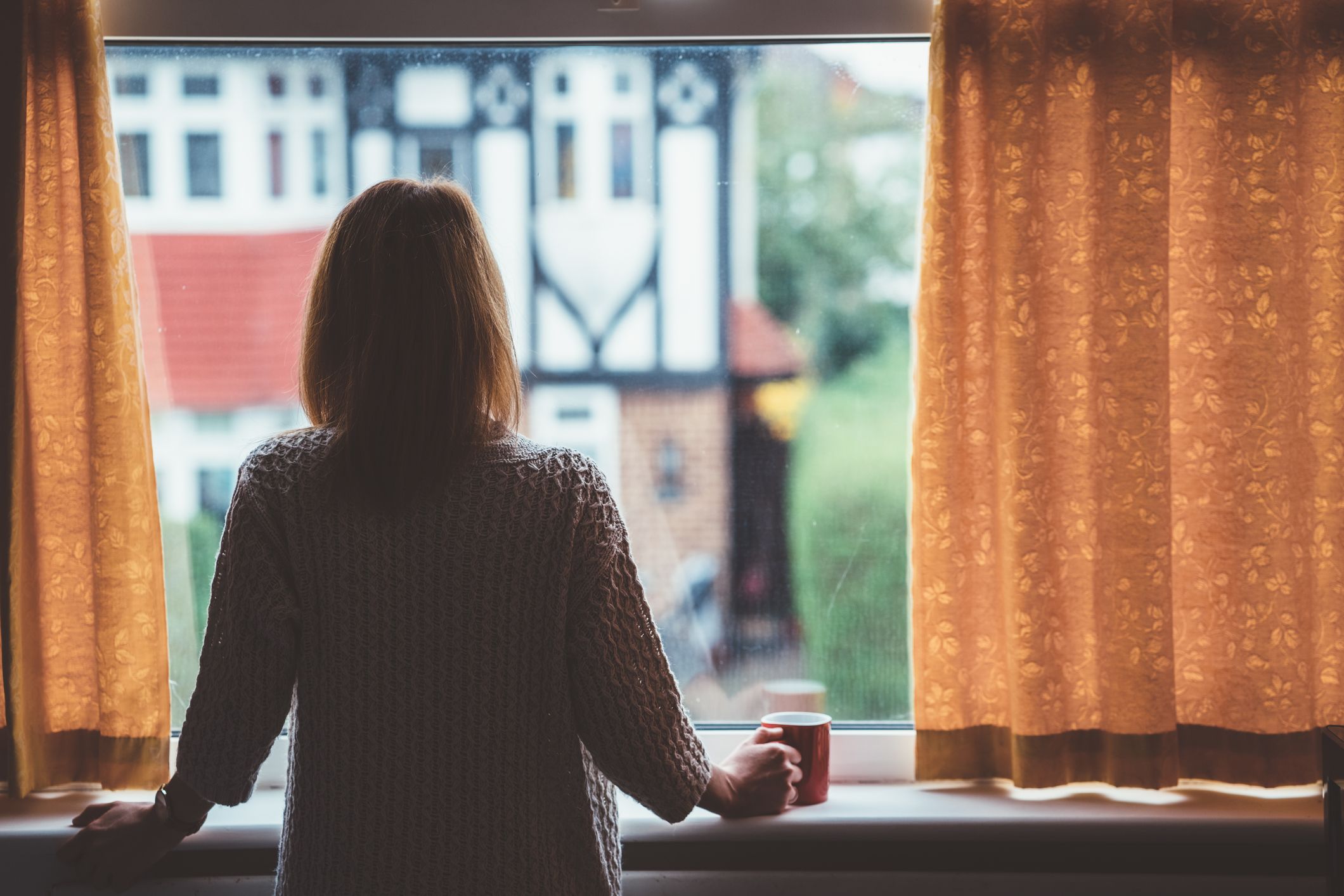 A middle aged woman stands looking out of her window