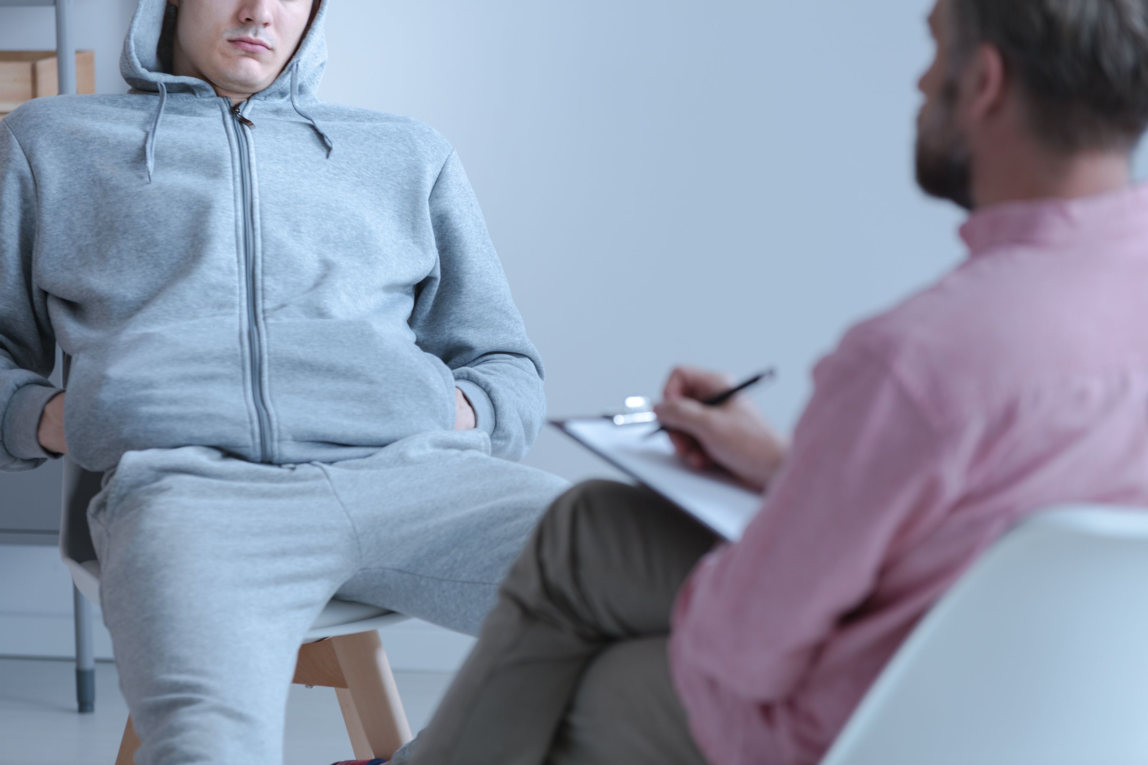 A young person wearing grey joggers and a grey hoodie speaks to a man taking notes on a clipboard